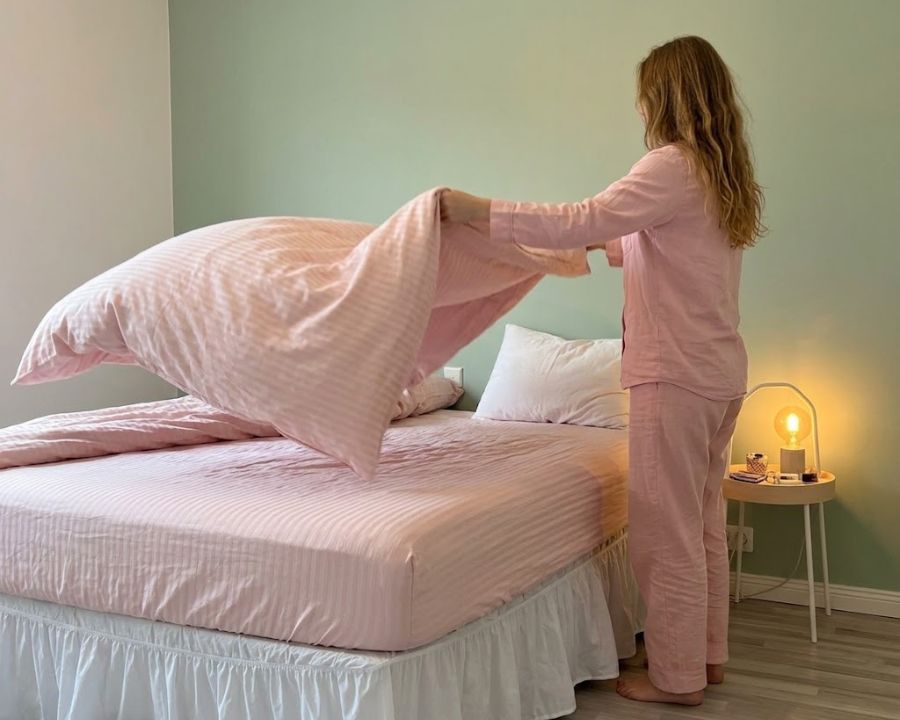 Good sleep hygiene habits of a girl making the sheets on the bed in the bedroom with light material pink sheets, green wall, and bedside tabe with calming light